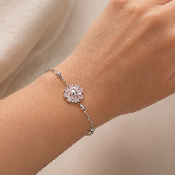 A delicate silver chain bracelet featuring a flower charm with light pink petals and a central clear stone, shown worn on a person's wrist against a soft beige background.