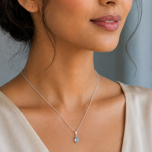 A close-up of a woman wearing a delicate silver chain necklace with a peardrop-shaped clear gemstone pendant, paired with a beige satin top.